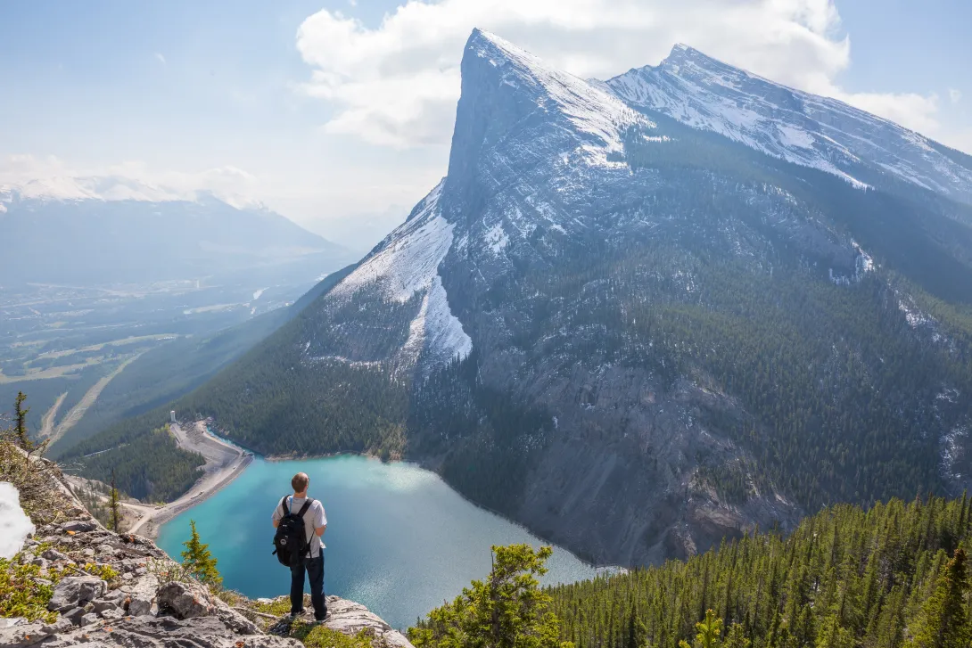 A backpacker standing on a high ledge looking out over a lake at the base of another mountain whose peak is covered in snow.