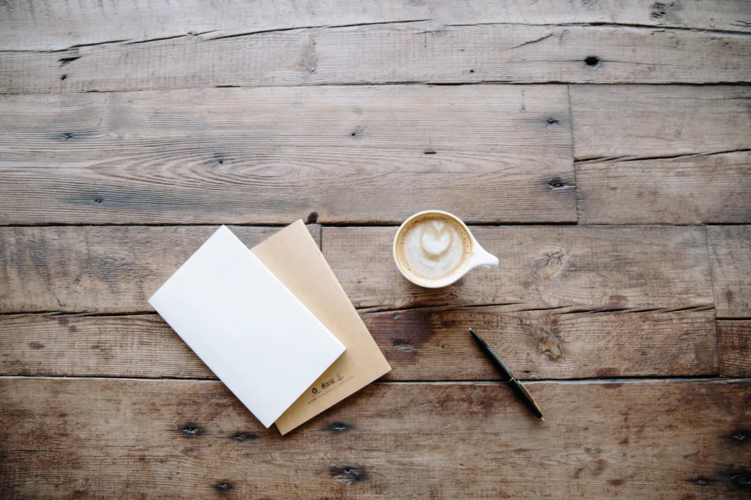 A picture of a table containing a notebook, pen and cup with a cappucino.